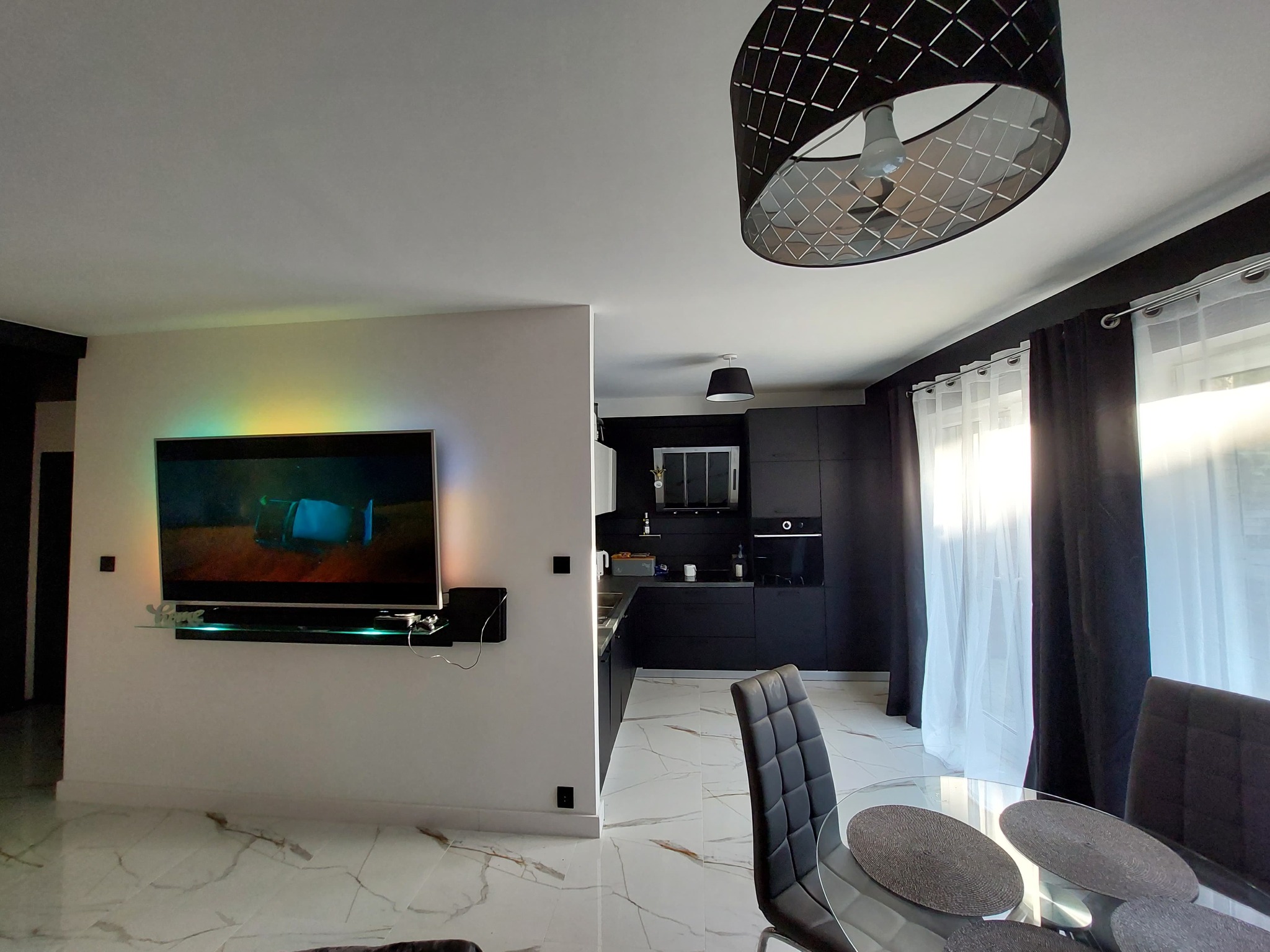 Nowoczesne wnętrze z marmurową podłogą, czarną kuchnią w tle i telewizorem podświetlonym kolorowym światłem na ścianie.