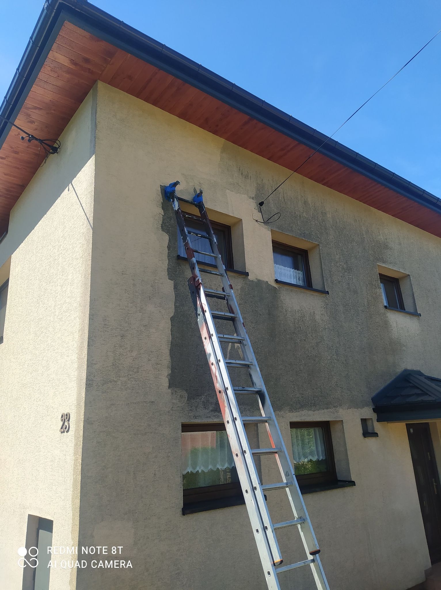 Malowanie elewacji domu jednorodzinnego z użyciem drabiny, widoczny fragment elewacji przed i po malowaniu, z numerem domu '23'.
