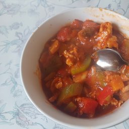 Ku zdrowiu - Słodko-kwaśne danie z kurczakiem, papryką i ananasem w białej misce, z łyżką zanurzoną w sosie, na stole z kwiatowym obrusem.