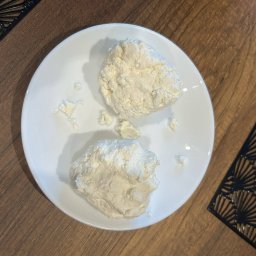 Ku zdrowiu - Dwie porcje twarogu na białym talerzu, ustawione na drewnianym stole z czarnymi, ażurowymi podkładkami. Delikatne światło podkreśla strukturę sera.