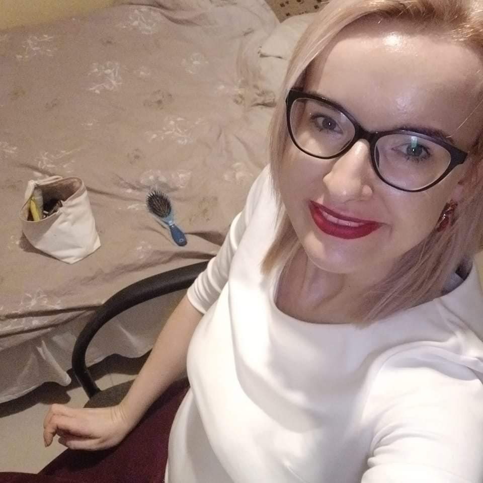 Portret kobiety w okularach i białej sukience, z kosmetyczką i szczotką do włosów na łóżku w tle.