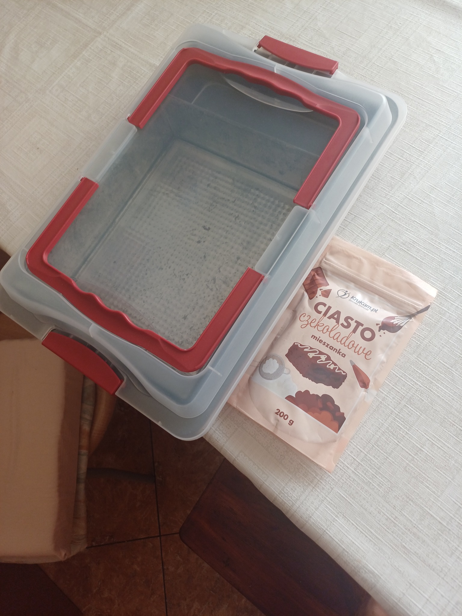 Pudełko transportowe na ciasto z czerwonymi uchwytami i paczka mieszanki do ciasta czekoladowego leżą na stole z jasnym obrusem.