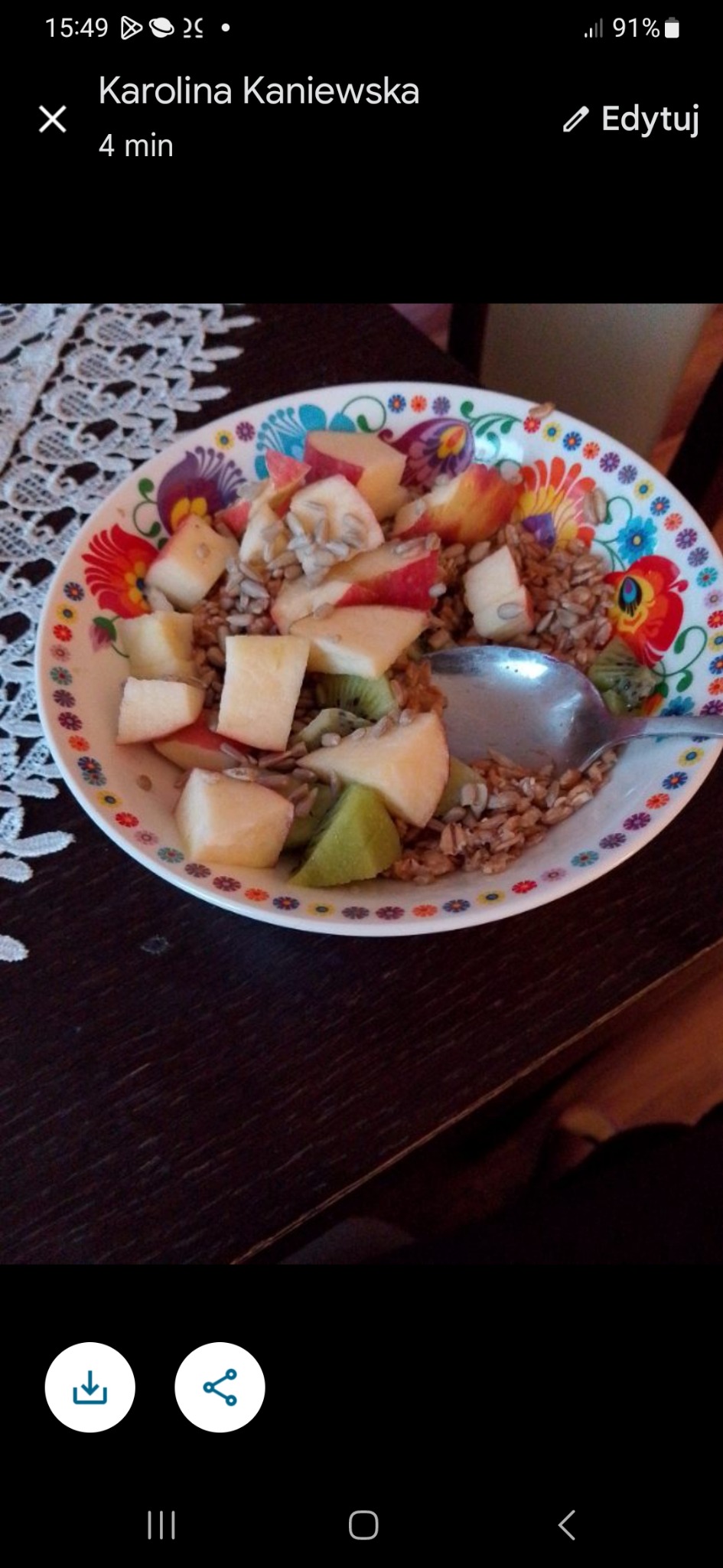 Kolorowy talerz z owsianką, jabłkami, kiwi i nasionami słonecznika, częściowo zjedzony, z łyżką w środku, na ciemnym stole z koronkową serwetą w tle.