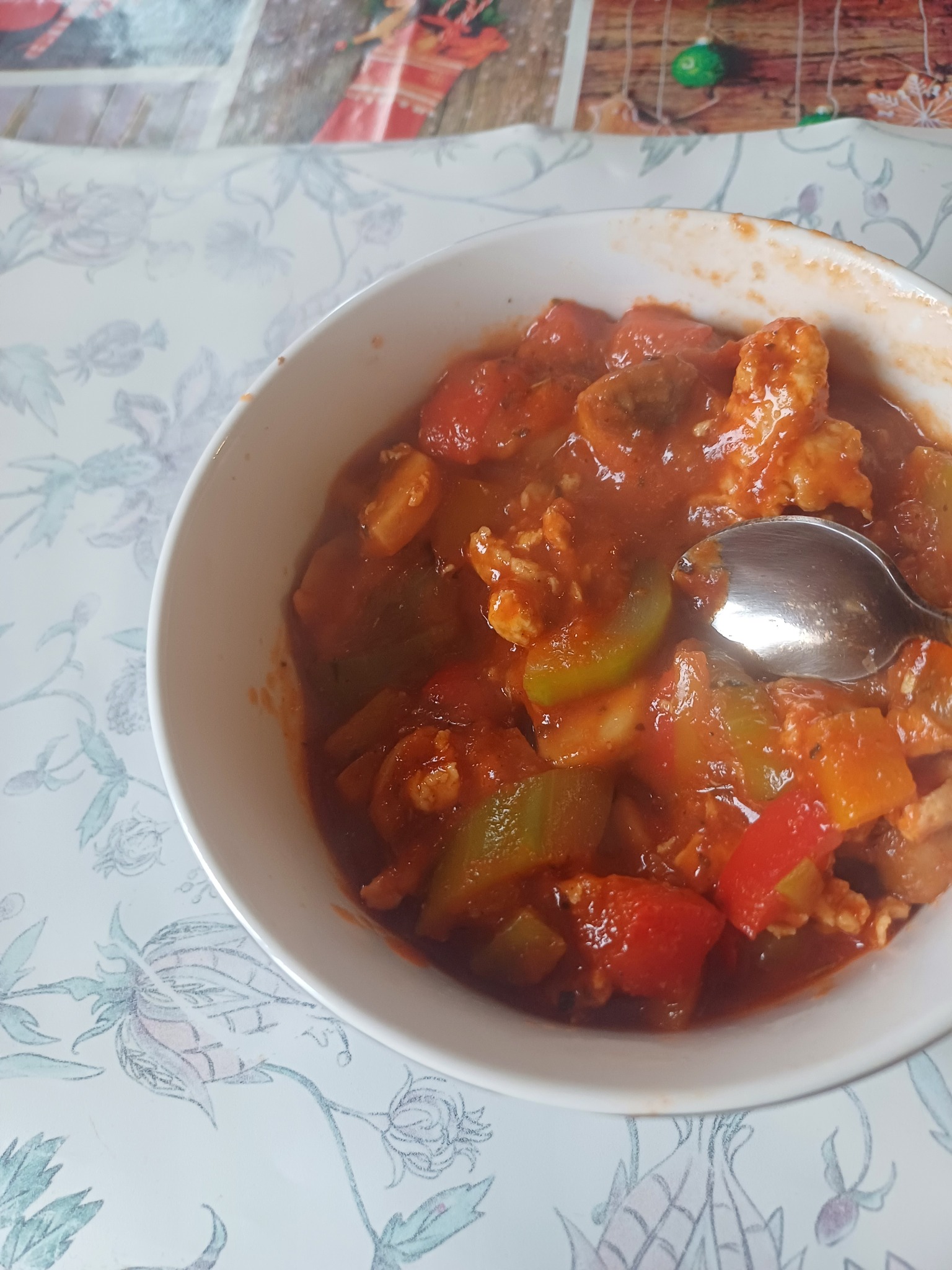 Słodko-kwaśne danie z kurczakiem, papryką i ananasem w białej misce, z łyżką zanurzoną w sosie, na stole z kwiatowym obrusem.