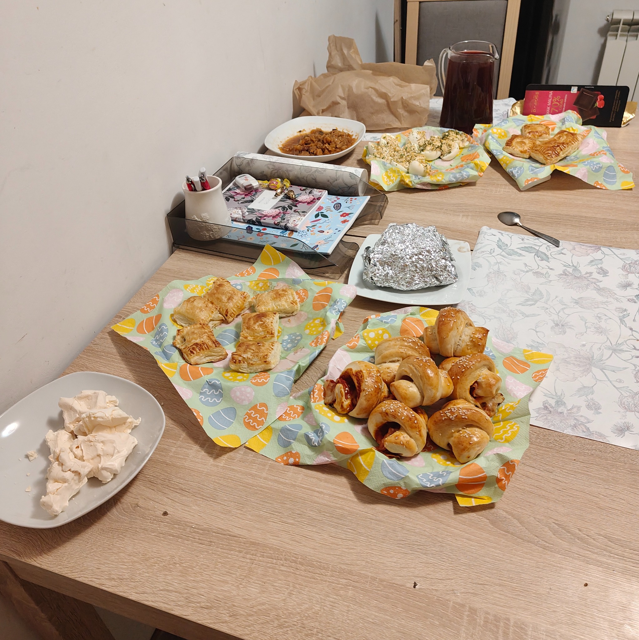 Stół zastawiony różnorodnymi potrawami: paszteciki z ciasta francuskiego, rogaliki z szynką posypane sezamem, jajka faszerowane, sałatka w sosie, dzbanek kompotu, czekolada, bezy na talerzu...