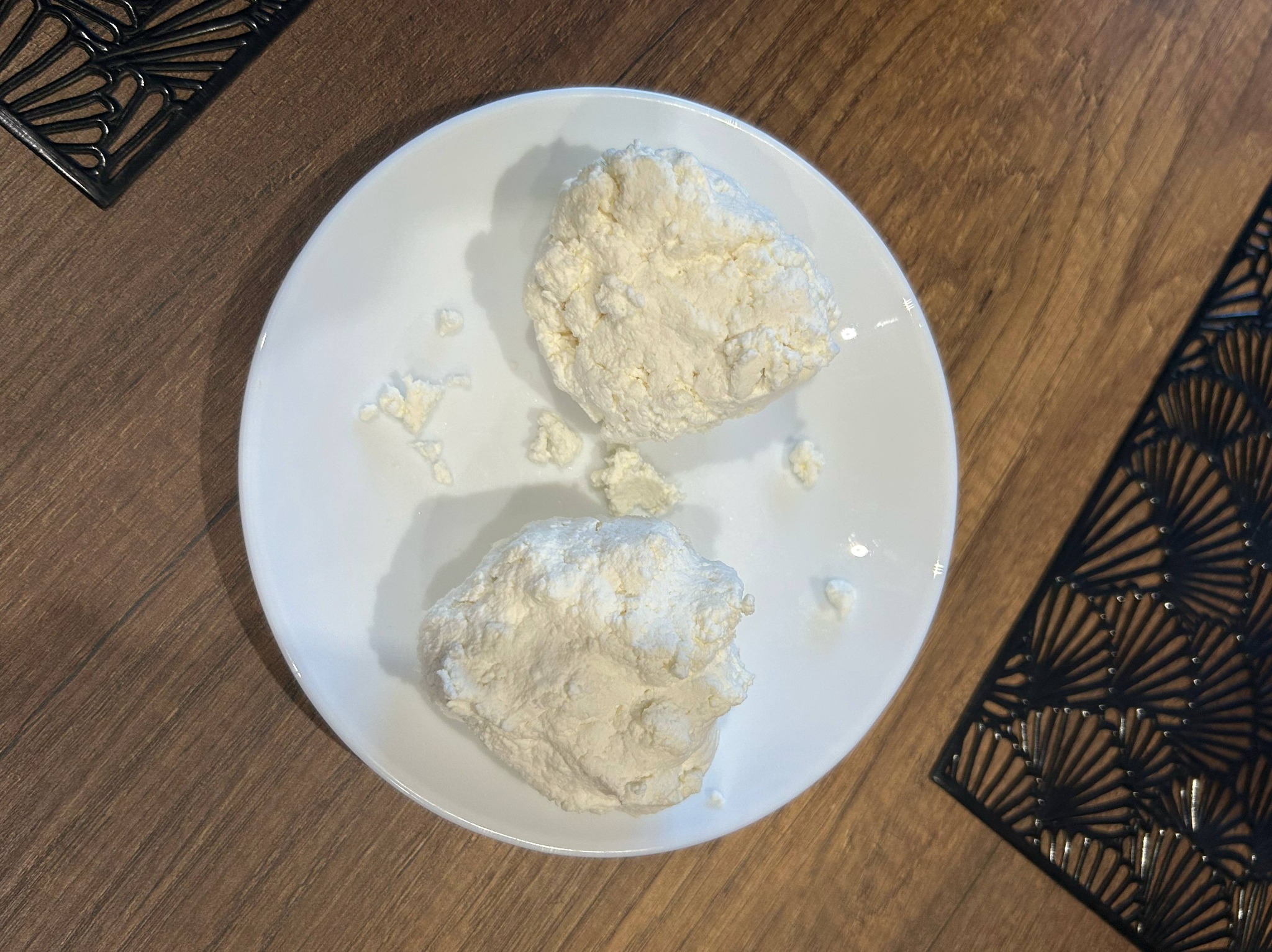 Dwie porcje twarogu na białym talerzu, ustawione na drewnianym stole z czarnymi, ażurowymi podkładkami. Delikatne światło podkreśla strukturę sera.