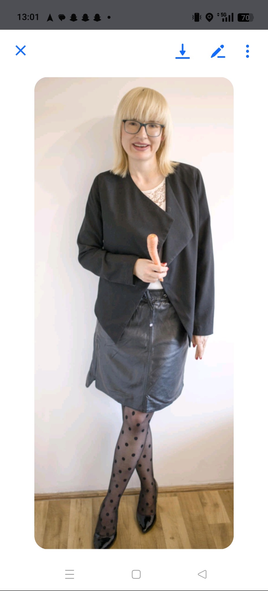 Dietetyczka w okularach trzyma marchewkę, ubrana w czarną marynarkę i skórzaną spódnicę. Rajstopy w kropki i czarne szpilki uzupełniają stylizację. Jasne tło.