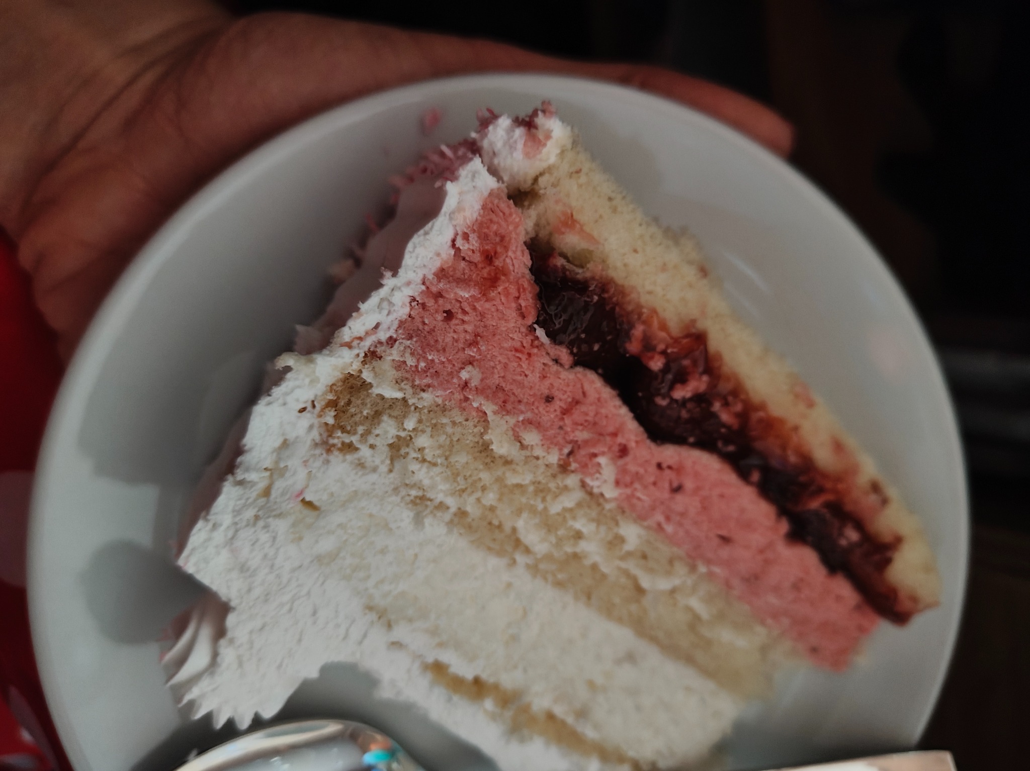 Kawałek tortu z kremem, różowym musem i dżemem na białym talerzu, trzymany w dłoni. Widoczne warstwy biszkoptu i kremu. Słodki deser z bliska.