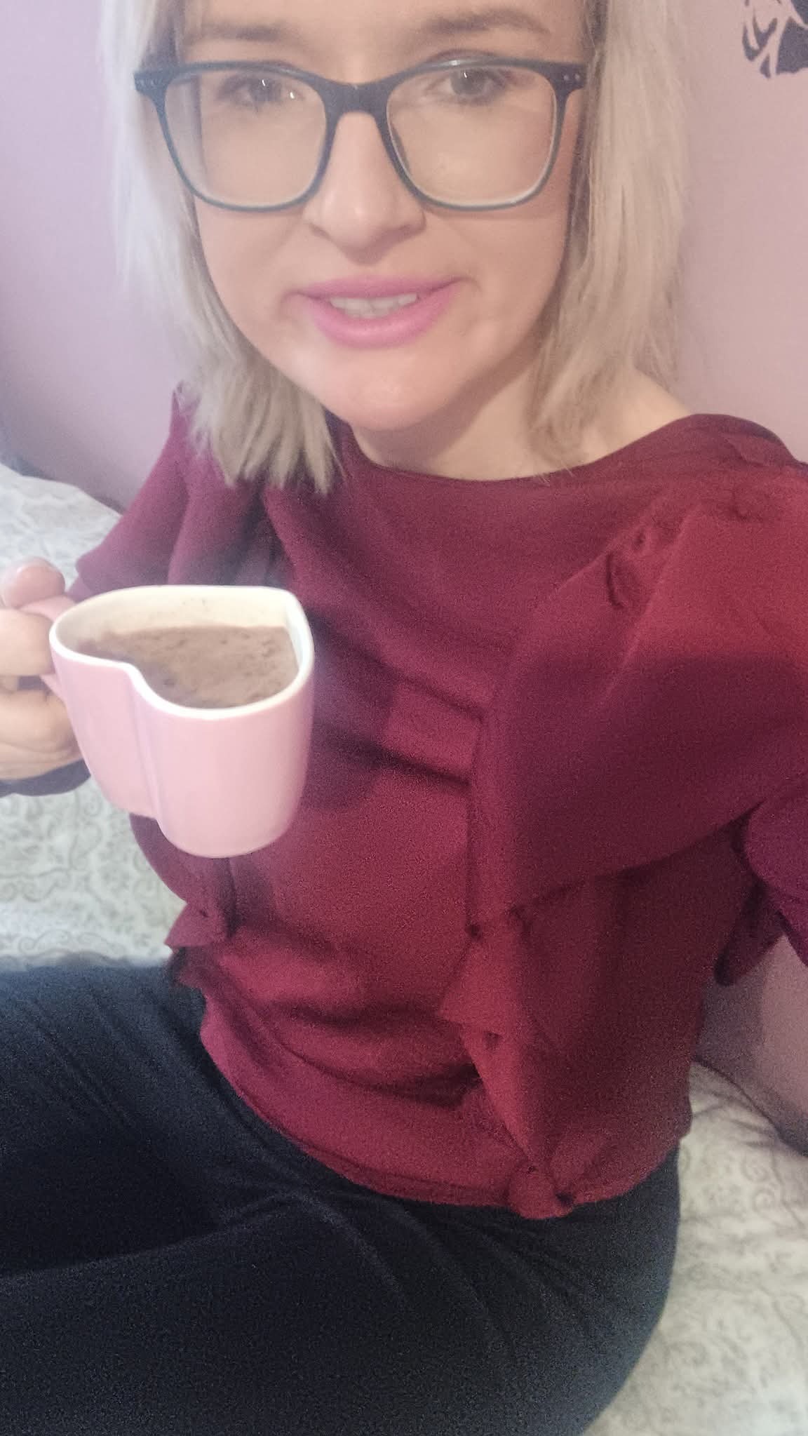 Kobieta w okularach pije z różowego kubka w kształcie serca. Widoczna bluzka w kolorze burgund i fragment wzorzystej pościeli. Uśmiech na twarzy.