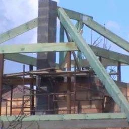 Wznoszenie konstrukcji dachu z zielonych belek drewnianych na murowanym budynku, widoczne rusztowanie i komin z bloczków betonowych, pochmurne niebo w tle.