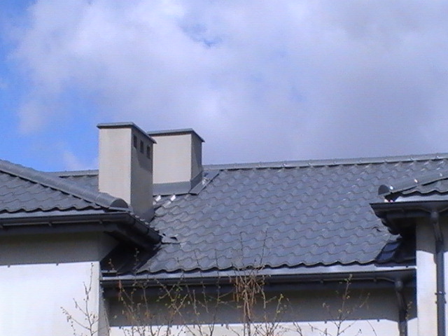 Szary dach z dachówki ceramicznej, dwa kominy z szarymi obudowami, orynnowanie w kolorze dachu, widoczne fragmenty białej ściany budynku, błękitne niebo w tle.