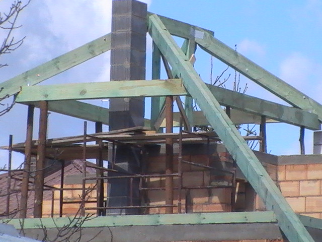 Wznoszenie konstrukcji dachu z zielonych belek drewnianych na murowanym budynku, widoczne rusztowanie i komin z bloczków betonowych, pochmurne niebo w tle.