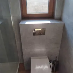 Nowoczesna toaleta podwieszana z szarą okładziną, drewnianym oknem z widokiem na zewnątrz i drewnopodobną podłogą, widoczny dozownik i szczotka do WC.