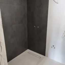 Fragment kabiny prysznicowej z ciemnoszarą glazurą na ścianach i jasnoszarą podłogą z odpływem liniowym, widoczne przygotowane punkty oświetleniowe i wyprowadzone kable elektryczne.