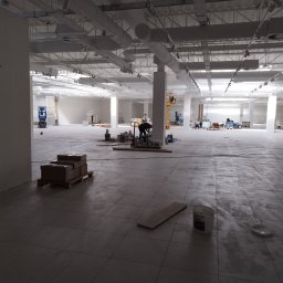 U.R. Rem- Max - Rozległa, jasna przestrzeń biurowa w trakcie remontu, z widocznymi pracownikami na podnośniku, materiałami budowlanymi i ułożoną częściowo podłogą z płytek. W tle widoczne instalacje sufitowe...