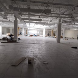 U.R. Rem- Max - Przestronne, jasne wnętrze biurowe w trakcie remontu, widoczne instalacje pod sufitem, pracownicy w oddali, podłoga w szarych płytkach, wiadro farby Brilux na pierwszym planie.