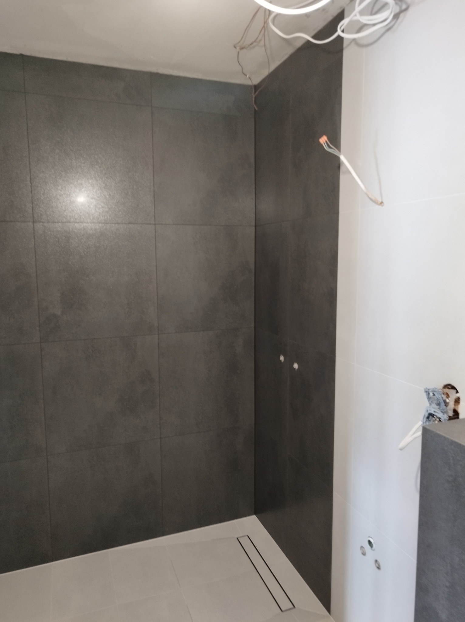 Częściowo wykończona łazienka z ciemnoszarymi płytkami na ścianach, widoczne niedokończone instalacje elektryczne i odpływ liniowy w podłodze.