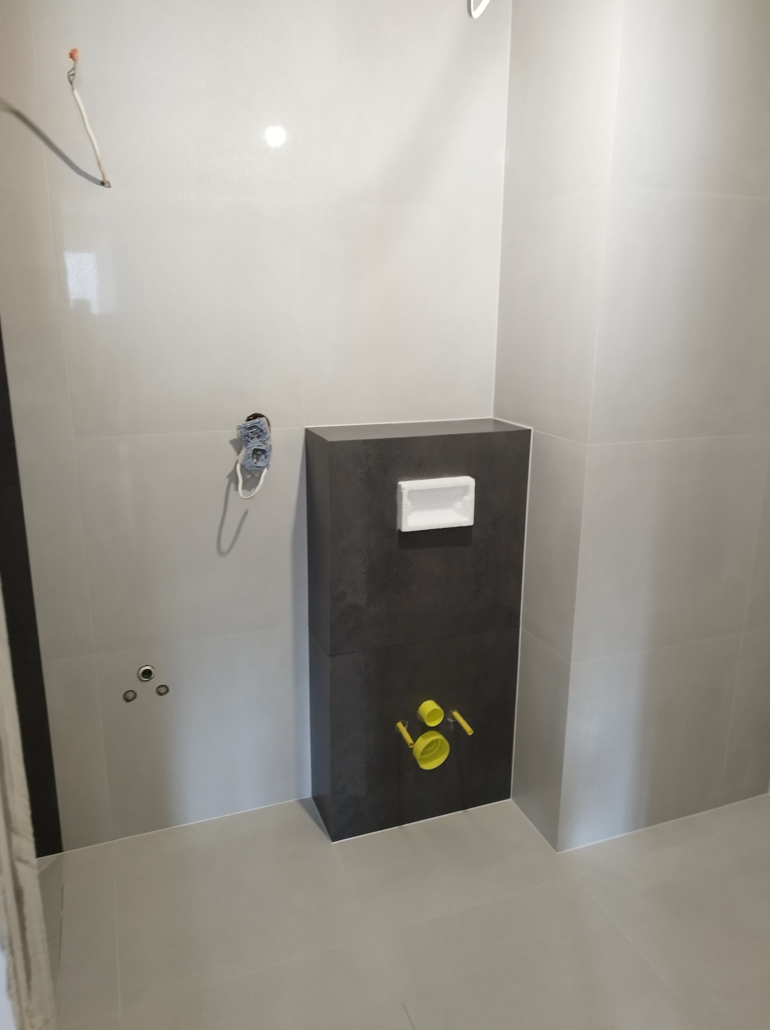 Szara łazienka w trakcie wykańczania, widoczna zabudowa stelaża WC z przyłączami, surowe płytki ścienne i podłogowe, wystające kable elektryczne, brak armatury.