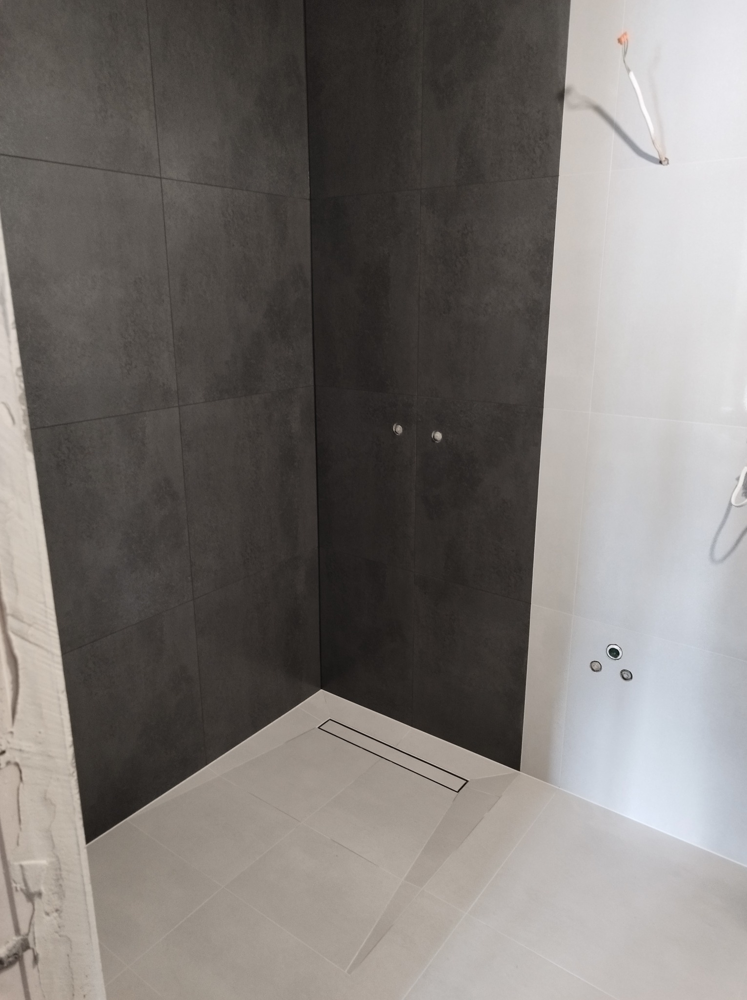 Fragment kabiny prysznicowej z ciemnoszarą glazurą na ścianach i jasnoszarą podłogą z odpływem liniowym, widoczne przygotowane punkty oświetleniowe i wyprowadzone kable elektryczne.