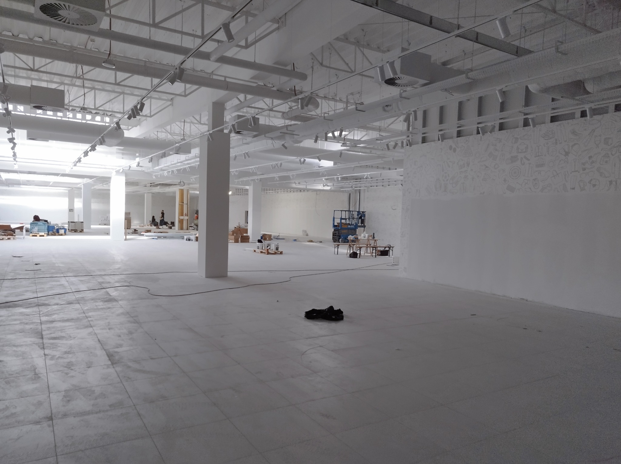 Przestronne wnętrze biurowe w trakcie remontu, widoczna surowa podłoga z płytek, białe ściany, instalacje pod sufitem, podnośnik i narzędzia w tle, przygotowania do wykończenia.