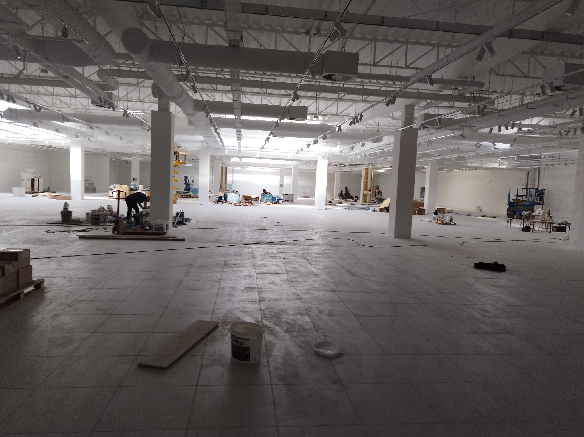 Przestronne, jasne wnętrze biurowe w trakcie remontu, widoczne instalacje pod sufitem, pracownicy w oddali, podłoga w szarych płytkach, wiadro farby Brilux na pierwszym planie.