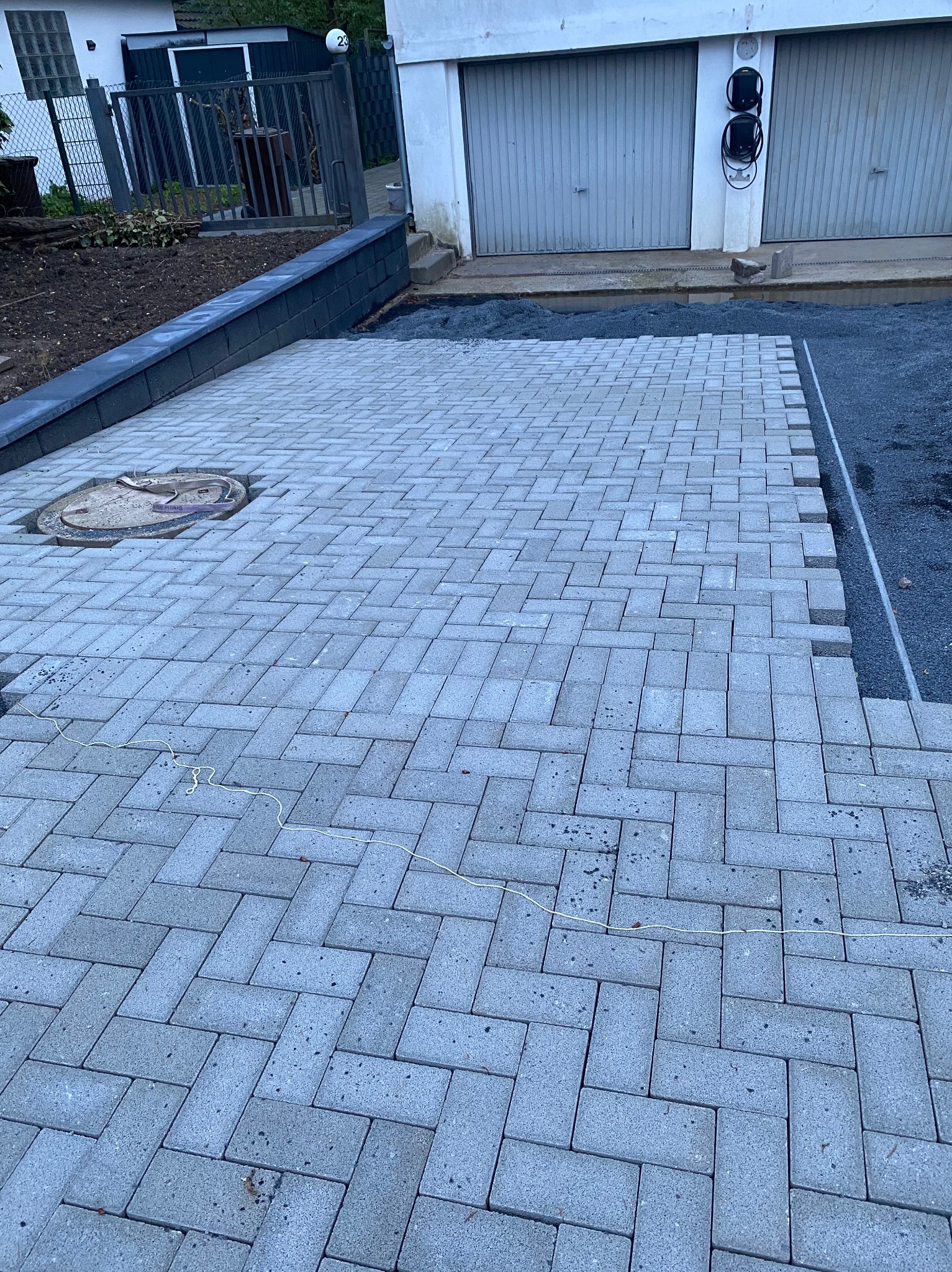 Fragment nowo ułożonej, szarej kostki brukowej w jodełkę, z widocznym włazem kanalizacyjnym i przygotowanym podłożem z boku, na tle garażu i fragmentu ogrodu.