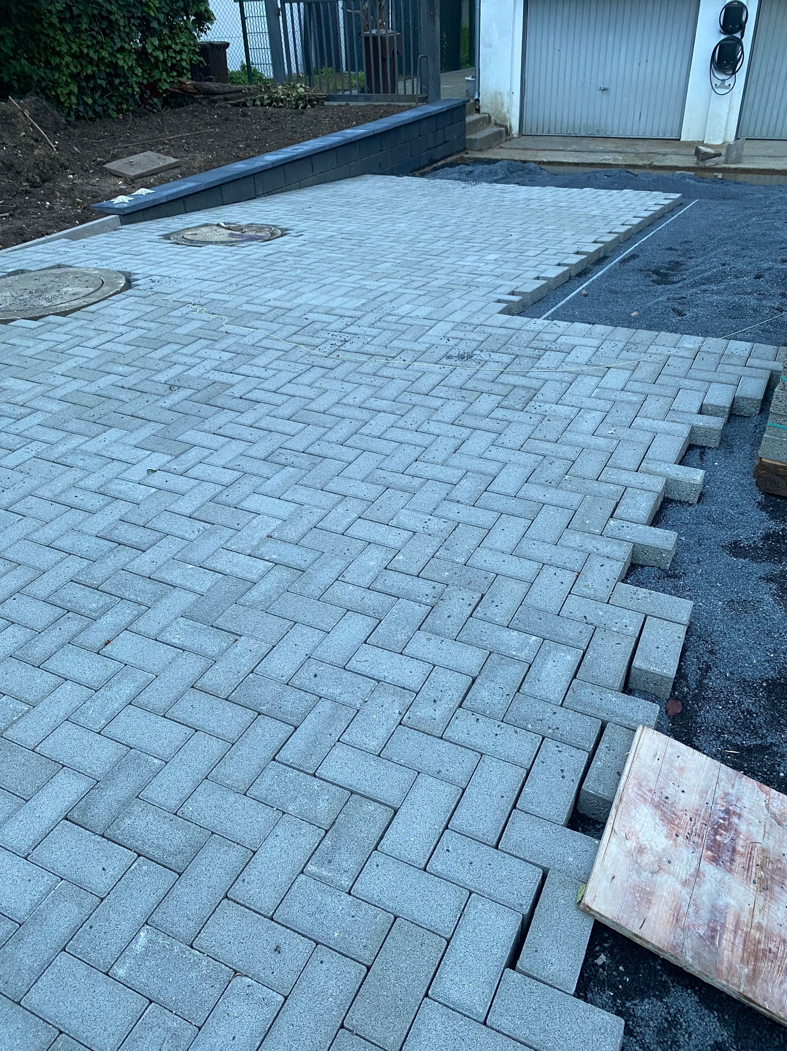 Układanie szarej kostki brukowej w jodełkę, widoczny fragment podsypki, studzienka kanalizacyjna i fragment garażu w tle.