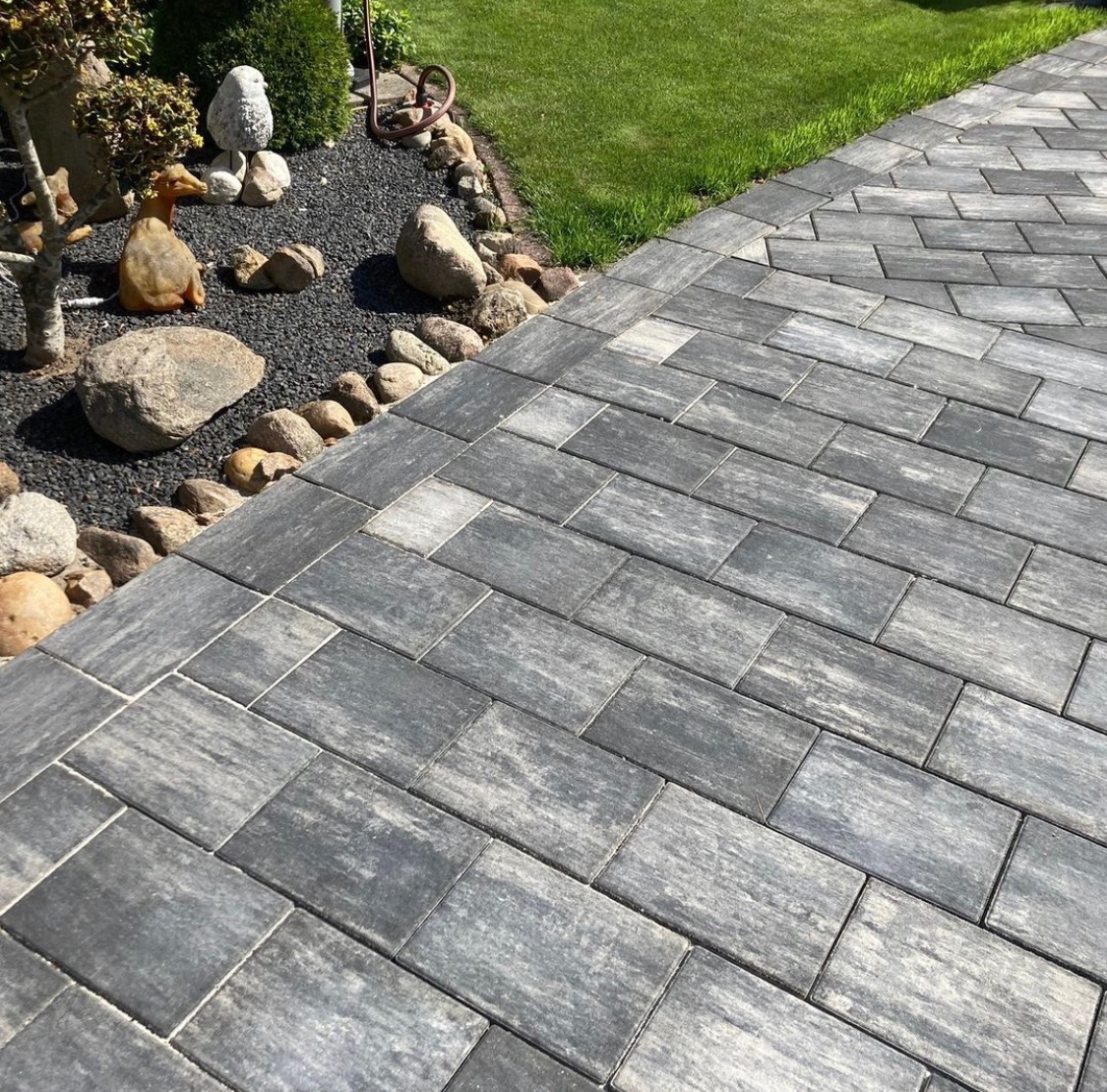 Szara, prostokątna nawierzchnia ułożona z kostki brukowej, obrzeża wykończone kamieniami, w tle trawnik i ozdobne krzewy.