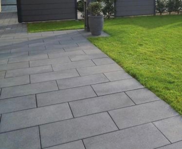 Szara nawierzchnia z prostokątnych płyt ułożonych w regularny wzór, obok zielony trawnik i ciemnoszare ogrodzenie. Donica z rośliną w tle.