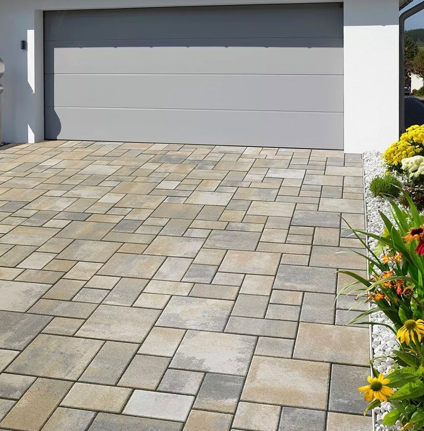 Ułożona kostka brukowa w odcieniach beżu i szarości prowadząca do garażu, obok rabata z kolorowymi kwiatami i białymi kamykami.