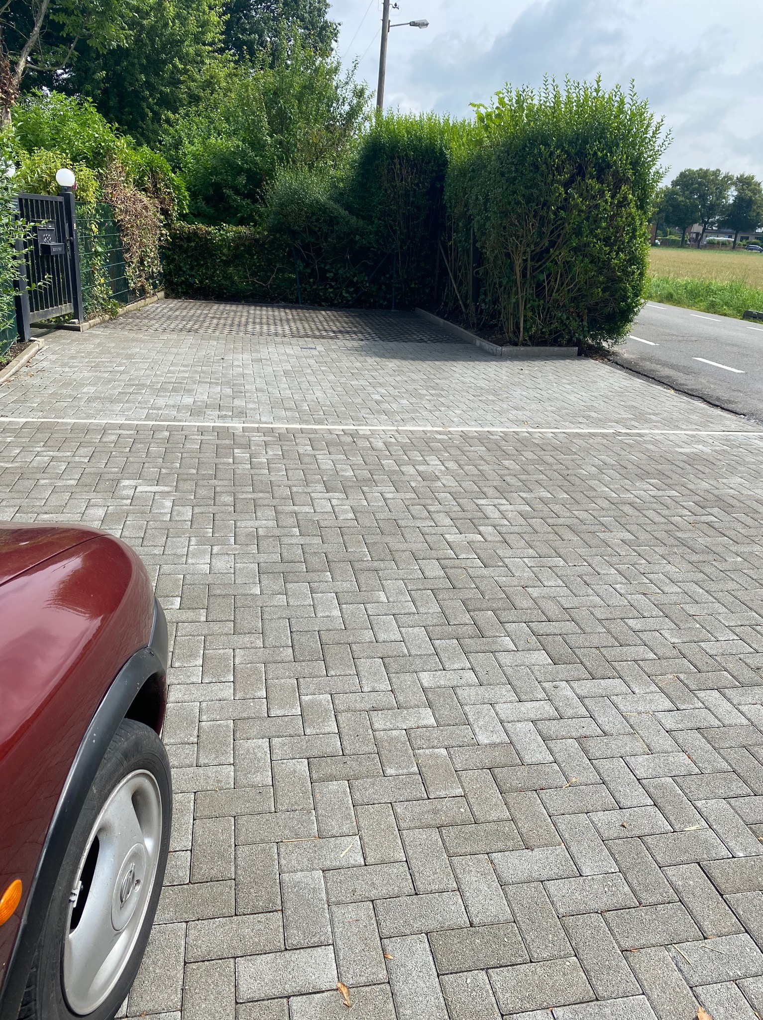 Nowo ułożona, szara kostka brukowa tworząca duży parking przed posesją, z fragmentem bordowego samochodu w lewym dolnym rogu i żywopłotem w tle.