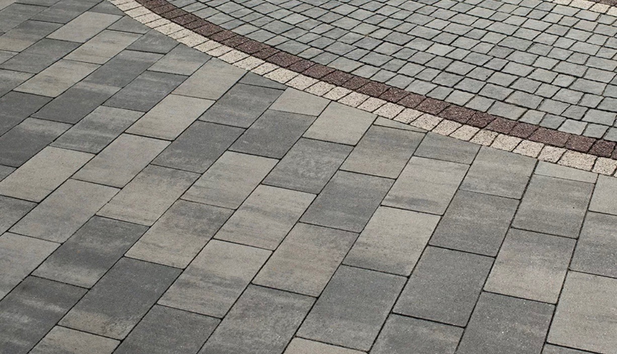 Ułożona nawierzchnia z prostokątnych szarych kostek brukowych ułożonych ukośnie, granicząca z obszarem wyłożonym drobną kostką w odcieniach szarości i brązu, tworząc geometryczny wzór.