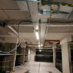 Metalowe korytka kablowe pod sufitem parkingu podziemnego, z widocznymi rurami, oświetleniem i strzałkami kierunkowymi na posadzce.