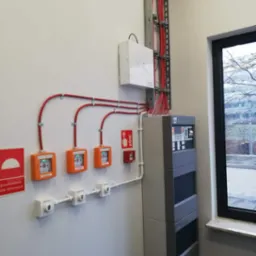 Centrala alarmowa z przyciskami oddymiania i ręcznym ostrzegaczem pożarowym na ścianie w budynku biurowym.