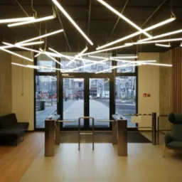 Nowoczesne oświetlenie biurowe:  system linearnych lamp LED zawieszonych pod sufitem, widoczne bramki wejściowe i fragment strefy oczekiwania z sofą i fotelem.