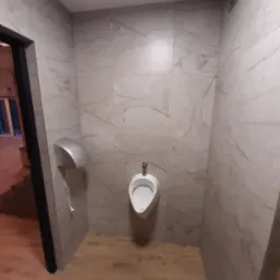 Umywalka pisuarowa zamontowana na ścianie z płytek imitujących marmur, obok suszarka do rąk, podłoga drewnopodobna, widok z perspektywy wejścia.