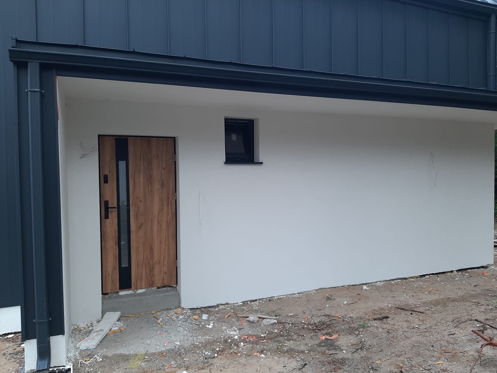 Wykończona elewacja budynku z zamontowanymi drzwiami wejściowymi z drewnopodobnym wzorem i małym oknem, widoczne orynnowanie i fragment terenu przed budynkiem.