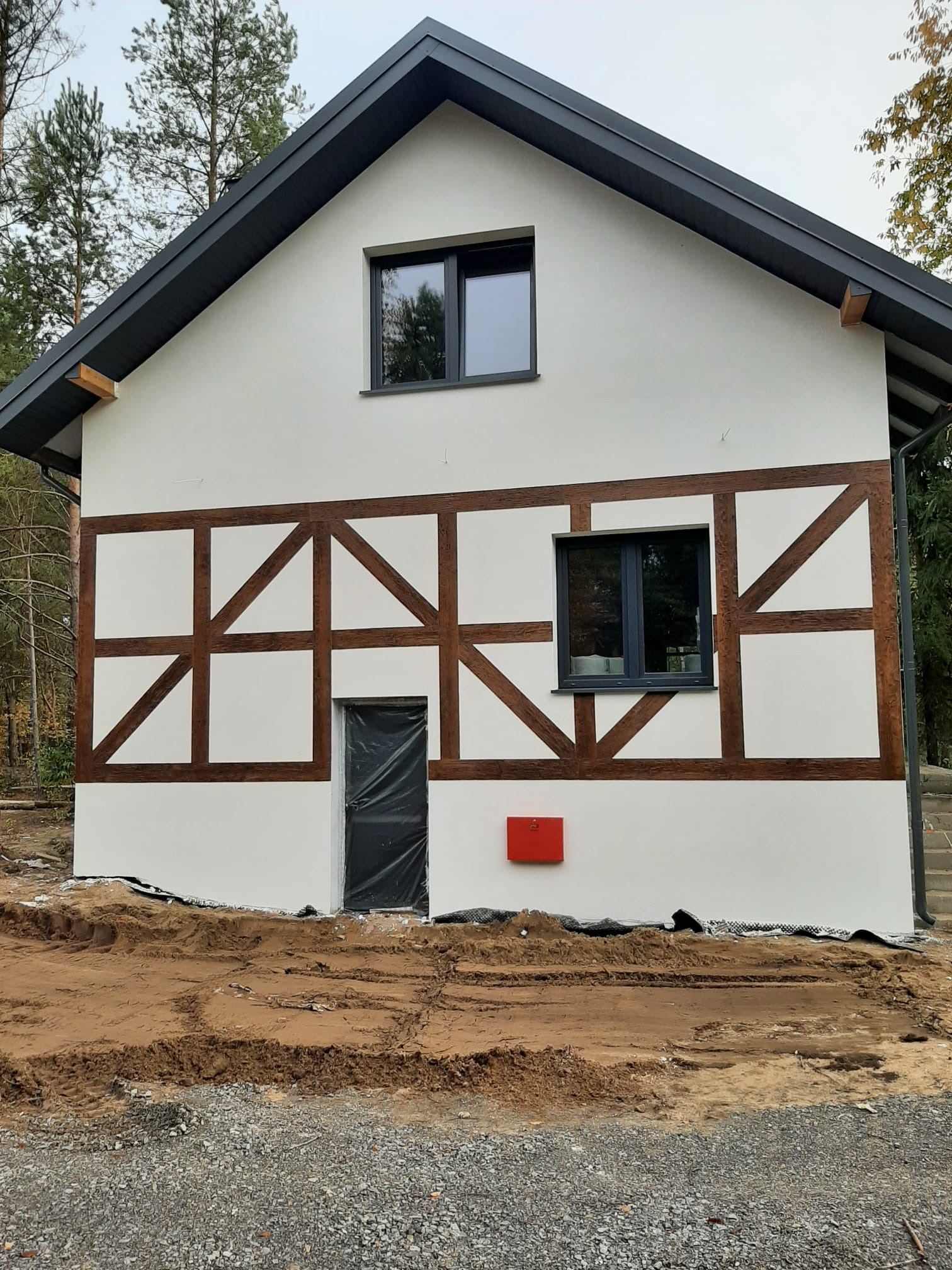 Biała elewacja domu z drewnianą konstrukcją szkieletową, czarnymi ramami okiennymi i tymczasowo zasłoniętymi drzwiami wejściowymi, otoczona ziemią przygotowaną pod zagospodarowanie terenu.