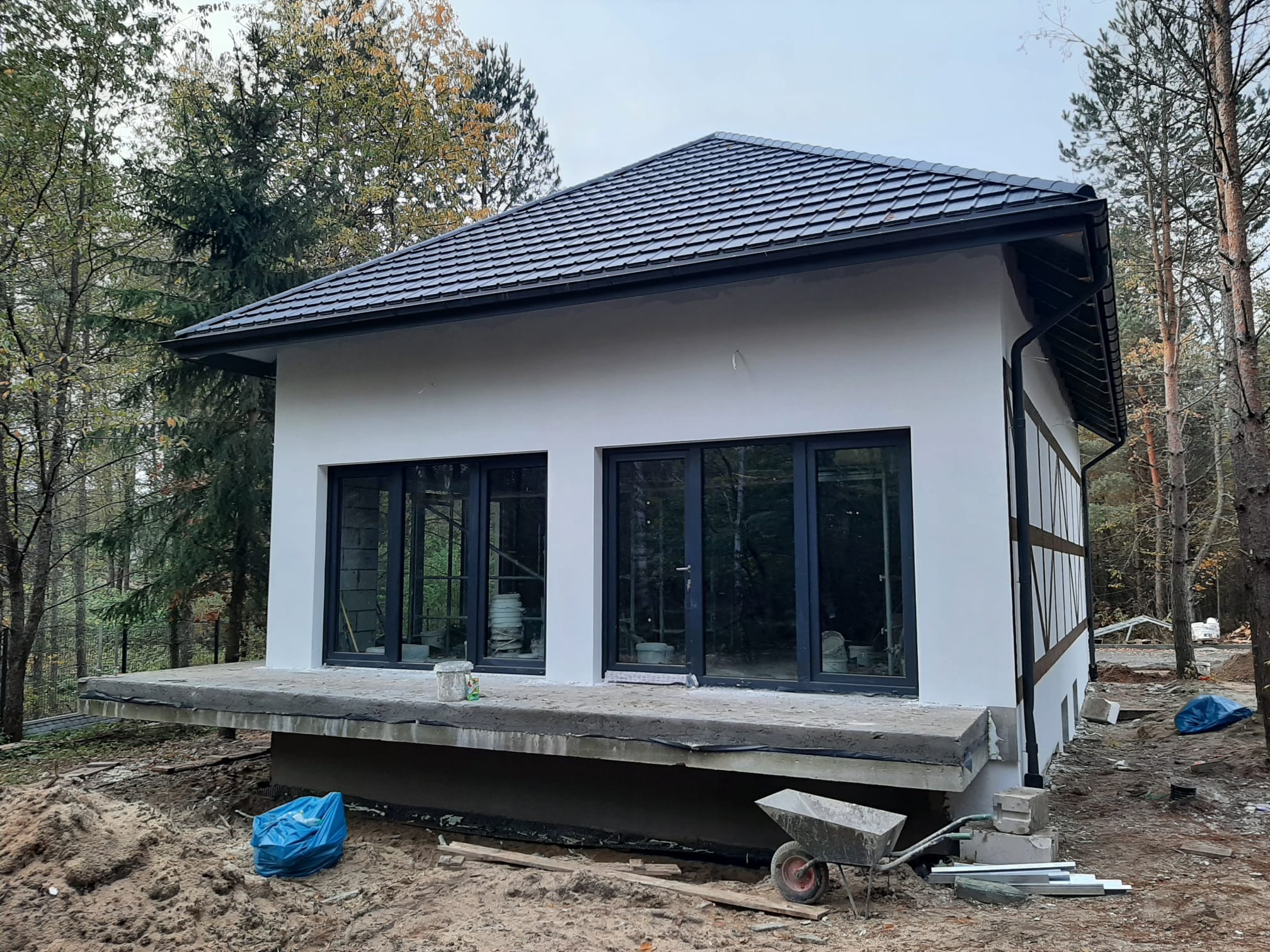 Niedokończony budynek z białymi ścianami i ciemnymi oknami, widoczna betonowa podstawa, narzędzia i otoczenie z drzewami.