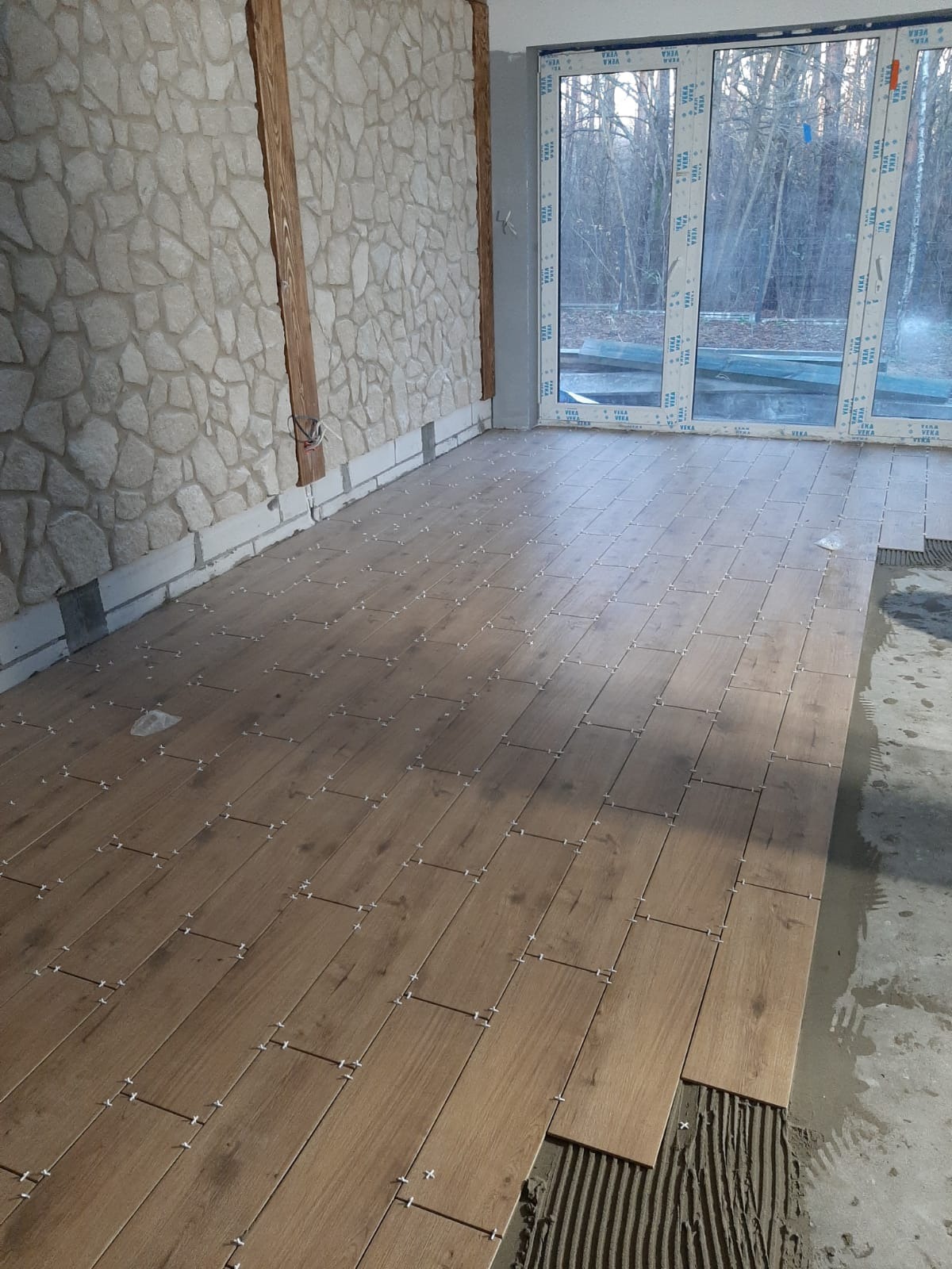 Układanie drewnopodobnych paneli podłogowych z białymi krzyżykami dystansowymi, widoczna ściana z kamienia dekoracyjnego i duże okno z widokiem na drzewa.