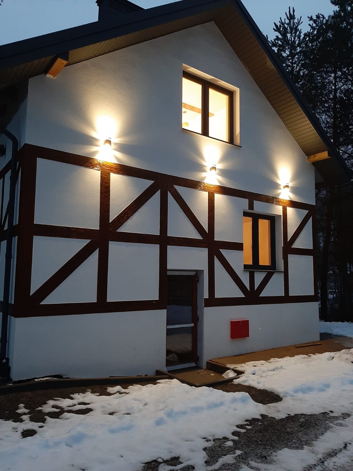 Oświetlona elewacja budynku w stylu szachulcowym z brązowymi belkami na białym tle, widoczne okna z ciepłym światłem oraz zewnętrzne lampy ścienne, fragmenty śniegu na ziemi.