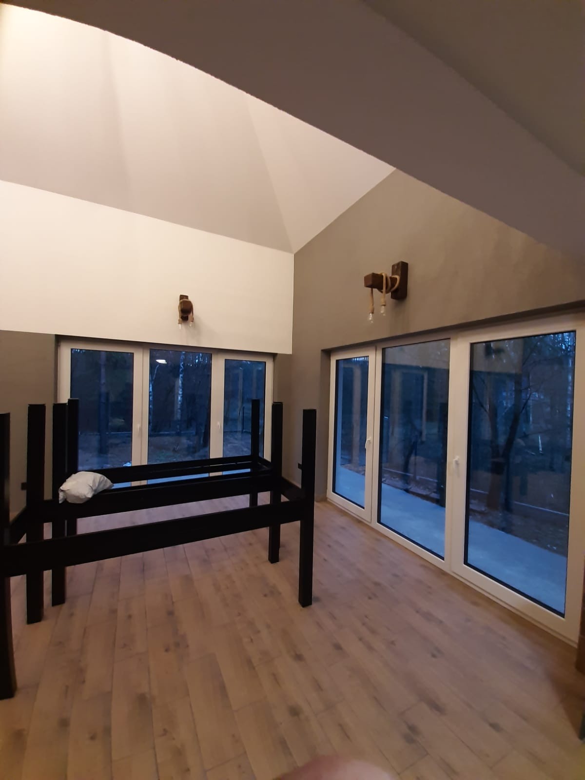 Jasne wnętrze z dużymi oknami balkonowymi i drewnianą podłogą. W centralnej części stoi czarna, metalowa konstrukcja przypominająca ramę łóżka lub stelaż meblowy, obok leży biała poduszka.