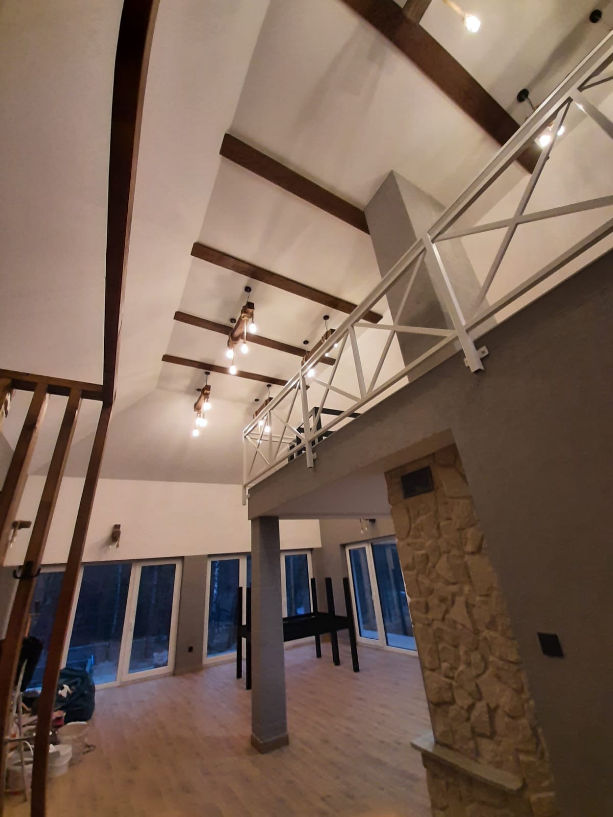 Przestronne wnętrze domu z antresolą, drewnianymi belkami stropowymi i dużymi oknami, widok z dołu na sufit i białą balustradę antresoli.
