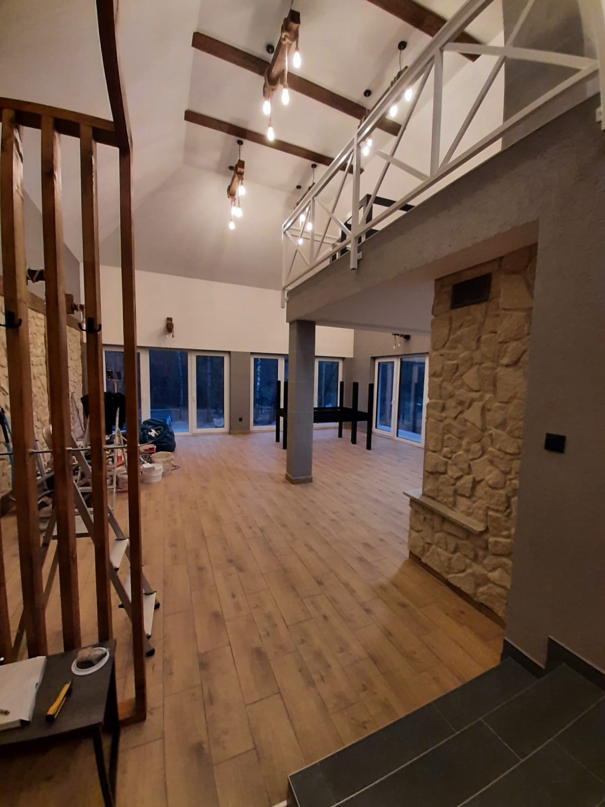 Przestronne wnętrze z drewnianą podłogą, widoczne schody z ciemnymi płytkami, kamienna ściana, balustrada na antresoli i surowe drewniane belki z żarówkami, w tle duże okna.