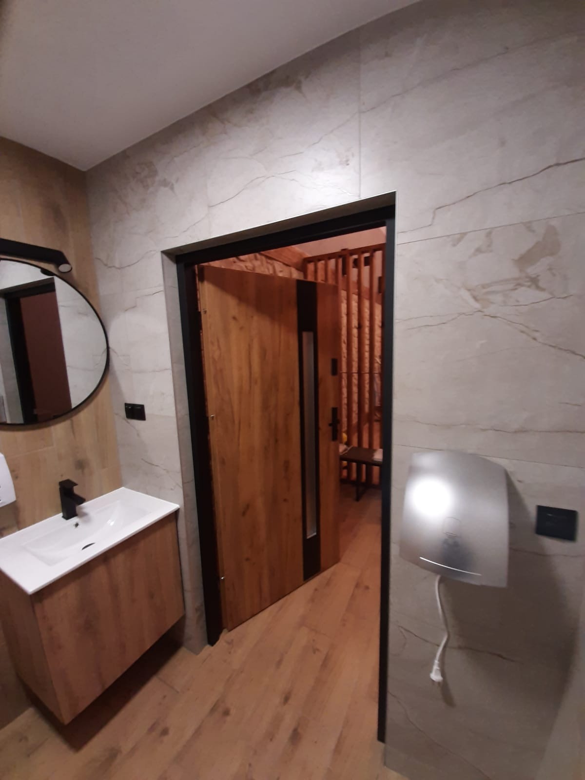 Nowoczesna łazienka z umywalką na szafce z drewnopodobnym fornirem, okrągłym lustrem z czarną obudową, suszarką do rąk i otwartymi drewnianymi drzwiami z czarnymi elementami prowadzącymi...