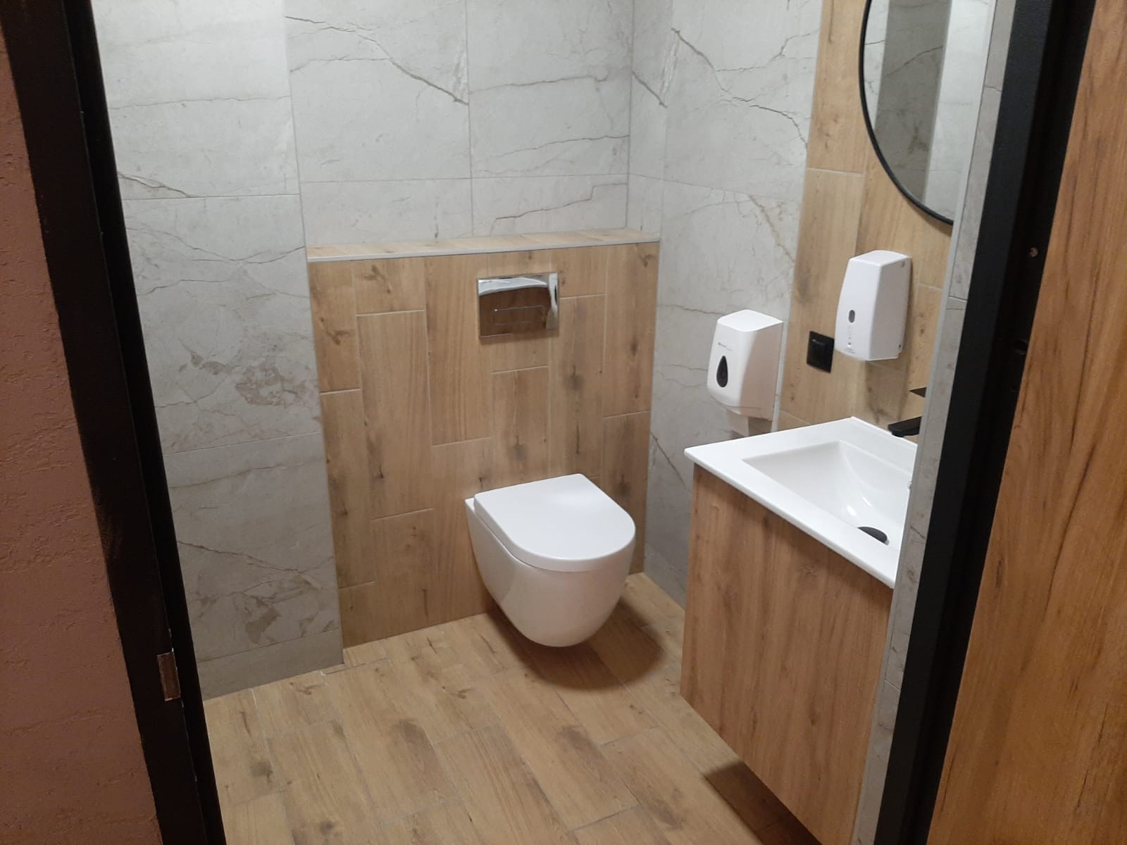 Nowoczesna łazienka z podwieszaną toaletą, umywalką z szafką w odcieniach drewna i dozownikami mydła na ścianie wyłożonej płytkami imitującymi marmur.