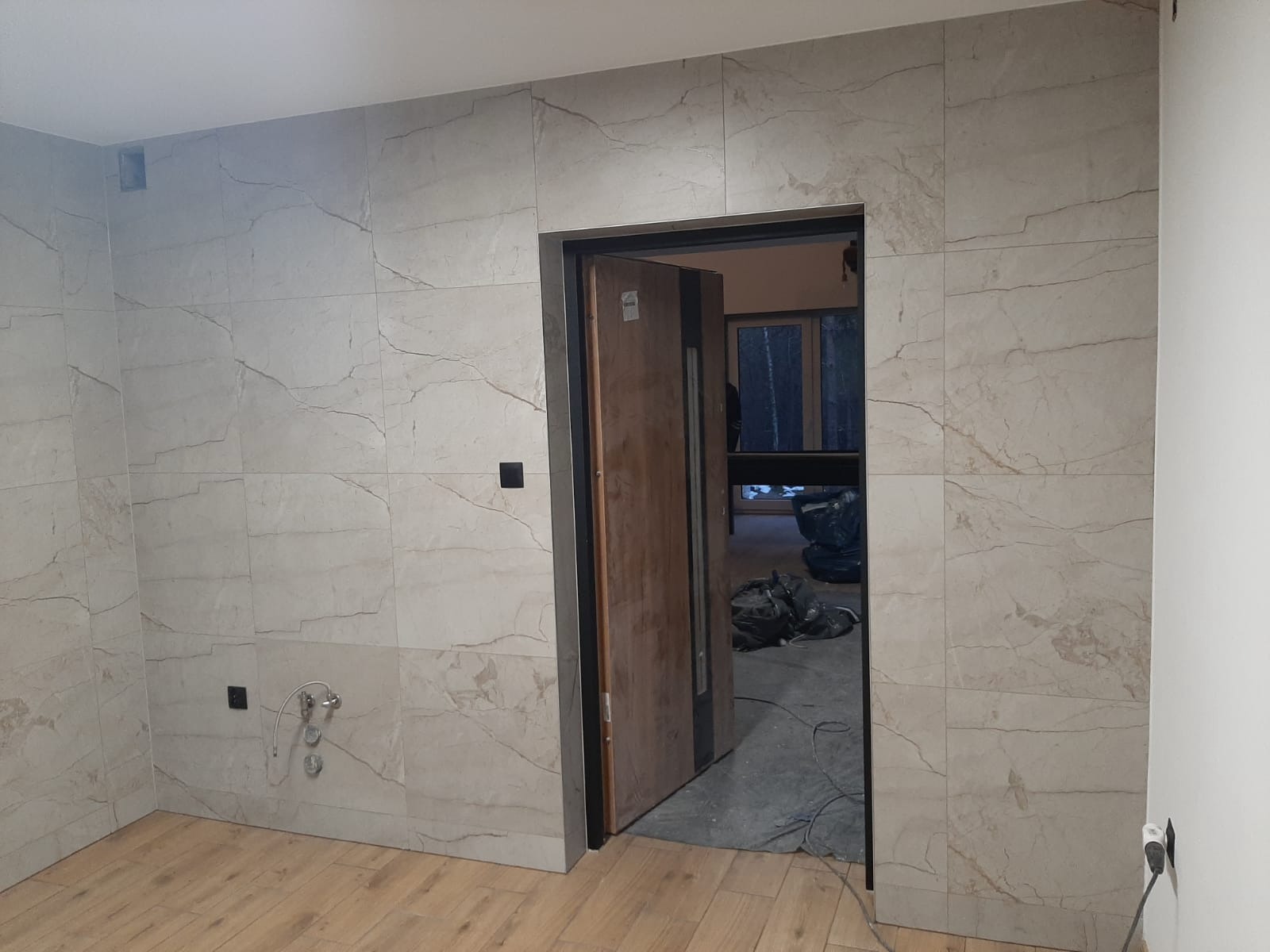 Wykończona łazienka z beżowymi płytkami imitującymi marmur na ścianach, drewnopodobnymi panelami na podłodze i otwartymi drzwiami z widokiem na remontowane wnętrze.