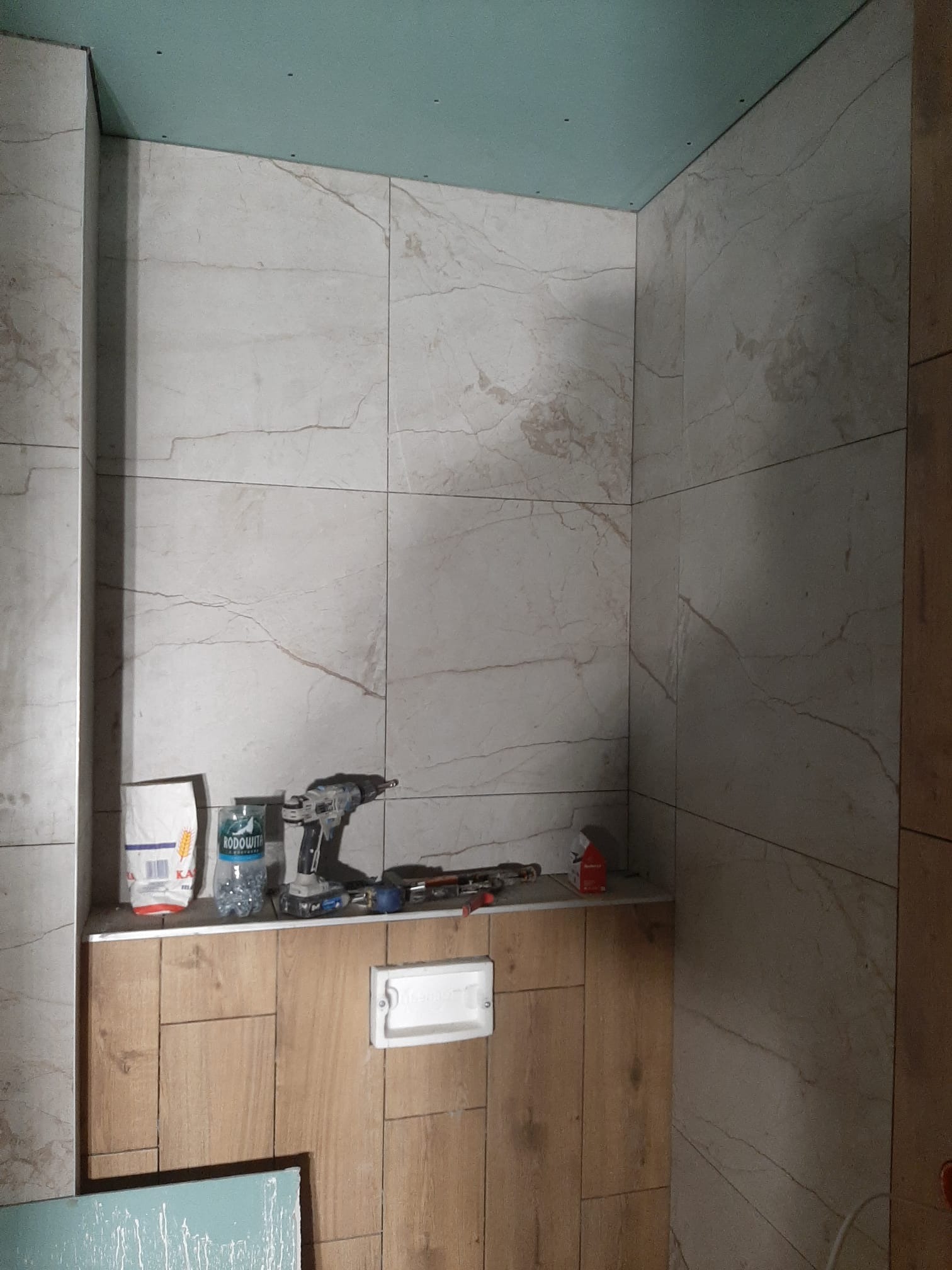 Wnętrze łazienki w trakcie remontu: ściany wyłożone płytkami imitującymi marmur, sufit z płyty gipsowo-kartonowej, widoczne narzędzia i materiały budowlane na półce.