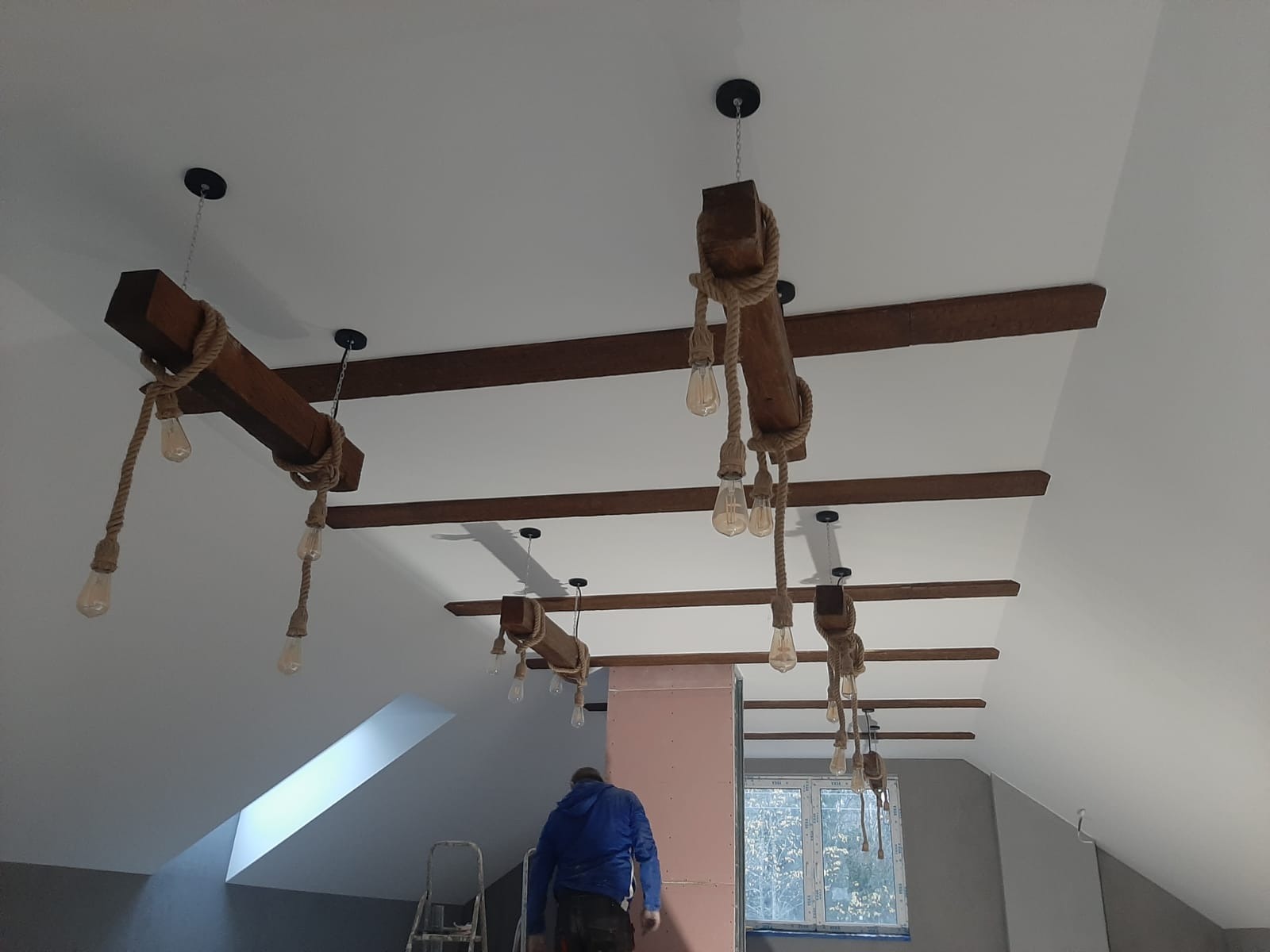 Montaż dekoracyjnych belek drewnianych z oświetleniem w pomieszczeniu ze skośnym sufitem, widoczna drabina i pracownik w niebieskiej bluzie.
