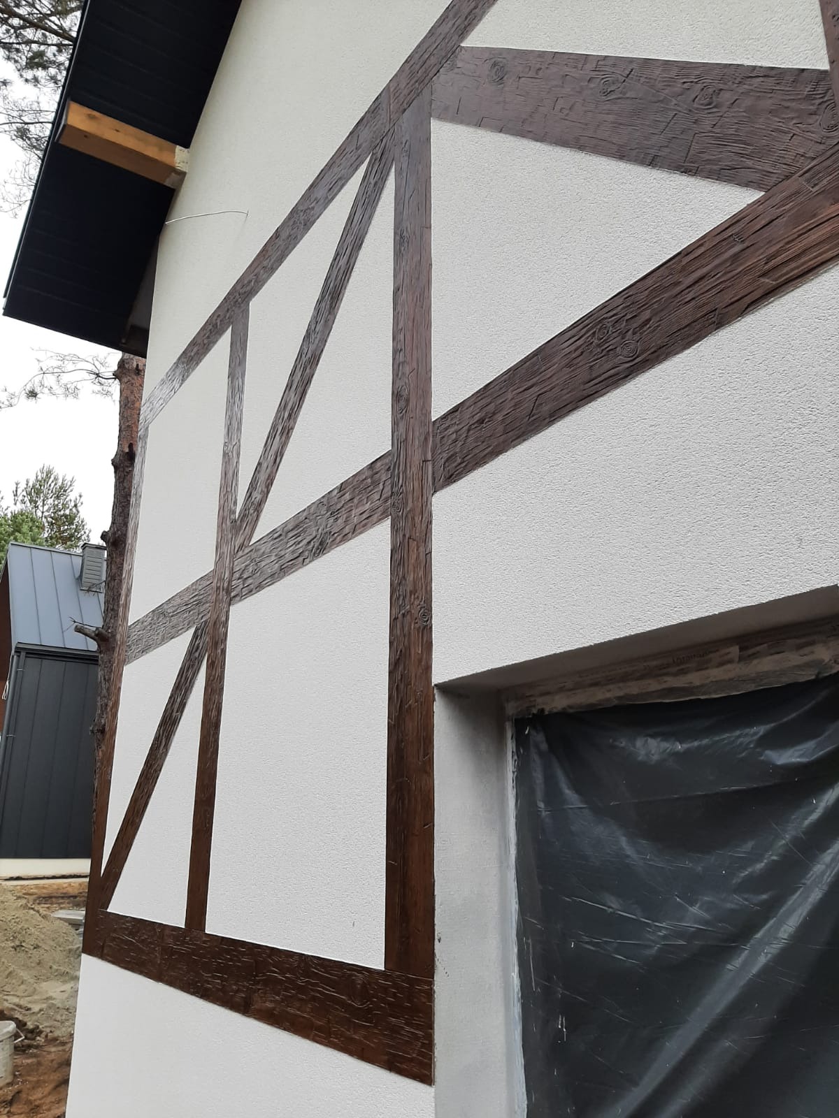 Fragment elewacji budynku z charakterystycznym wykończeniem w stylu muru pruskiego, z widocznym oknem zabezpieczonym folią ochronną.